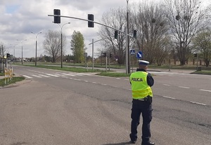 Policjant w kamizelce odblaskowej z napisem POLICJA stoi przy drodze w pobliżu przejścia dla pieszych i mierzy prędkość pojazdów